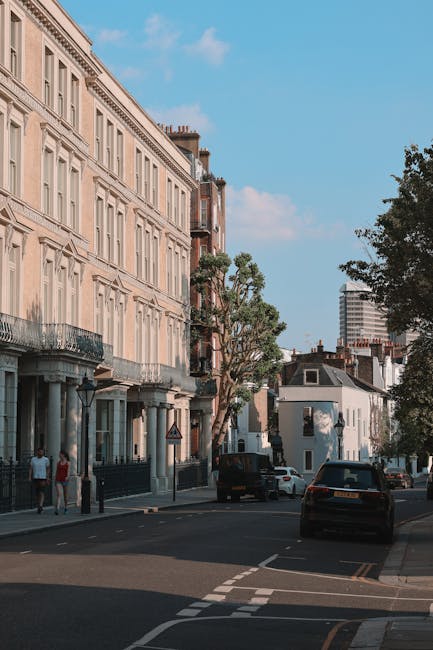 The image depicts a quiet residential street scene during daytime, featuring a row of multi-storey terraced houses with light-colored facades, decorative balconies, and large windows. In the foreground, there are several cardboard boxes and wrapped furniture pieces placed on the pavement, indicating a home relocation or packing process. A sturdy trolley is visible nearby, used for transporting packed items. Alongside the pavement, a black van is partially loaded with these boxes and furniture, with its rear doors open for access. A man from Man with Van Earls Court is seen loading boxes and furniture into the van, using straps and blankets to secure and protect the items during transport. The street is lined with trees and parked cars, and the weather appears clear with blue sky and some clouds, providing good lighting for the loading process. This scene illustrates the steps involved in furniture transport and packing during a house removal, with professional removal services supporting home relocation activities in London.