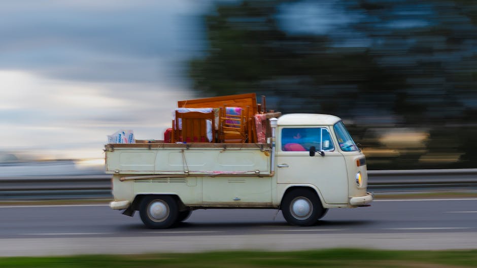 A vintage cream-colored van with a boxy shape is shown in motion on a roadway during daylight, carrying various household items and furniture in its open cargo area. Inside the cargo space, there are wooden chairs, a small table, and cardboard boxes, some wrapped in plastic or fabric. The van is being loaded or transported as part of a home relocation or furniture transport process, with items secured and arranged for safe transit. The background features blurred trees and sky, indicating the vehicle's movement, and the van's side mirror and windshield are visible, along with the vehicle's simple design typical of classic commercial vans. This scene illustrates the loading process associated with house removals, supported by the presence of a professional removals company, such as Man with Van Earls Court, specialized in packing and moving services.