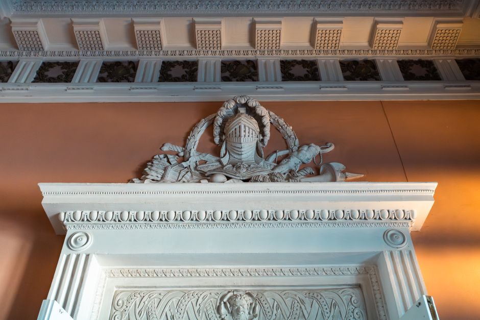 Inside a property, a detailed white decorative plaster relief featuring a helmeted figure, possibly a warrior, lying amidst various objects such as scrolls and weapons. The relief is mounted above a w